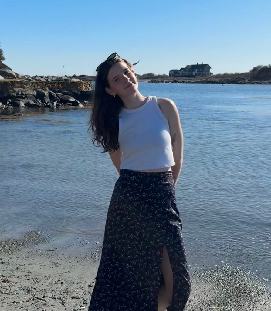 A person stands on a sandy shoreline beside calm blue water, tilting their head slightly and smiling softly at the camera. They are wearing a white sleeveless crop top and a black floral maxi skirt with a front slit, with sunglasses resting on their head. Rocky edges frame the water, and a large house sits across the inlet under a clear blue sky on a sunny day.