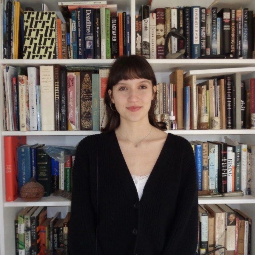 A person with dark hair and bangs stands in front of a large white bookshelf filled with books. They are wearing a black cardigan over a light-colored top, hoop earrings, and a necklace. The shelves behind them contain books of various sizes and colors arranged upright and stacked horizontally, creating a study or home library setting.