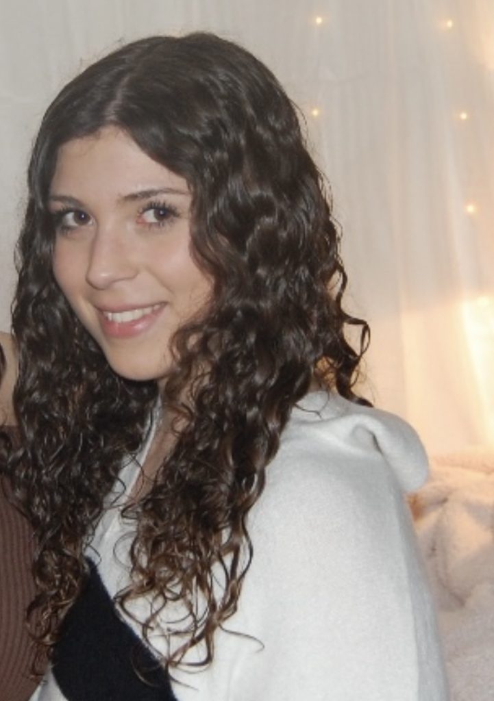 A close-up portrait of a smiling person with long, dark curly hair wearing a cream-colored sweater with navy stripes. They are looking slightly to the side while posing indoors in front of a soft white backdrop decorated with warm string lights. Part of another person is visible at the edge of the frame. The lighting creates a cozy, relaxed atmosphere.