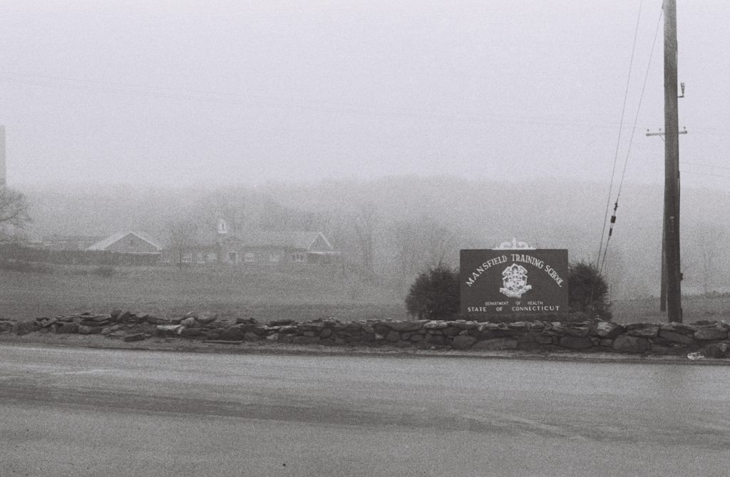 Black-and-white photograph of the entrance to Mansfield Training School on a foggy day. A stone wall runs along the roadside in the foreground, with a large sign reading "Mansfield Training School, Department of Health, State of Connecticut." Institutional buildings and leafless trees are faintly visible through the dense mist in the background, while utility poles and overhead wires frame the right side of the image.