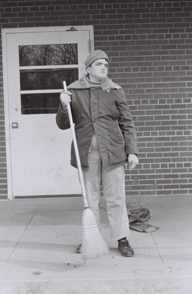 A person wearing a winter coat and knit hat sweeps an outdoor concrete walkway with a broom while standing in front of a brick building entrance. A pair of gloves rests on the ground nearby.