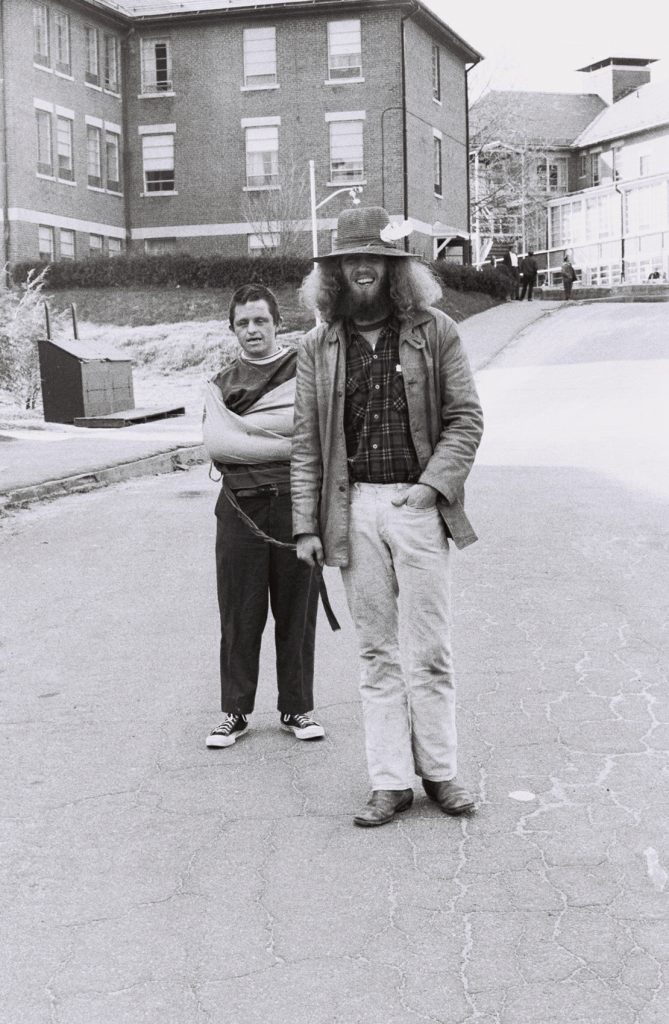 Black-and-white photograph of two people standing on a road between institutional buildings. One person smiles while wearing a wide-brimmed hat and long coat, while the other stands behind with one arm in a sling.
