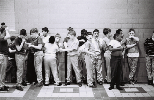Black-and-white photograph of a group of children standing along a cinder block wall in a tiled hallway. Several children appear to be interacting or jostling with one another, while others stand apart or look toward the camera. Some faces and bodies are blurred, suggesting movement.