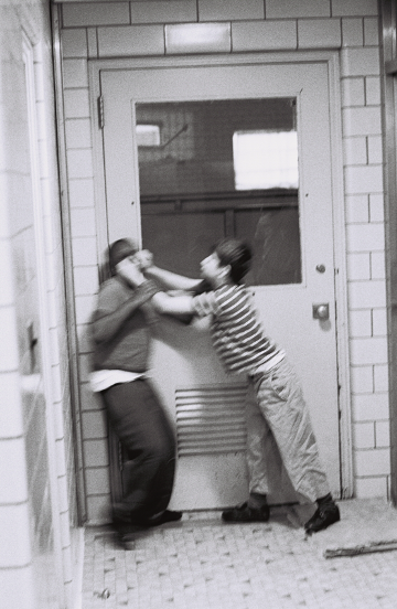 Black-and-white photograph of two children in a tiled hallway engaged in a physical altercation in front of a closed door. One child is pushing the other against the door with both hands, while the other leans back with their hands raised defensively. Motion blur emphasizes the struggle.