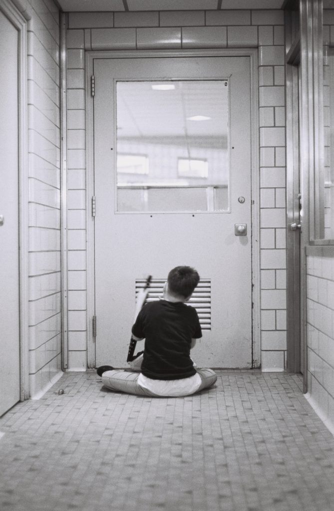 Black-and-white photograph of a person sitting cross-legged on a tiled floor facing a closed door. They hold a long object in one hand, and the hallway is lined with tiled walls and doors on either side.