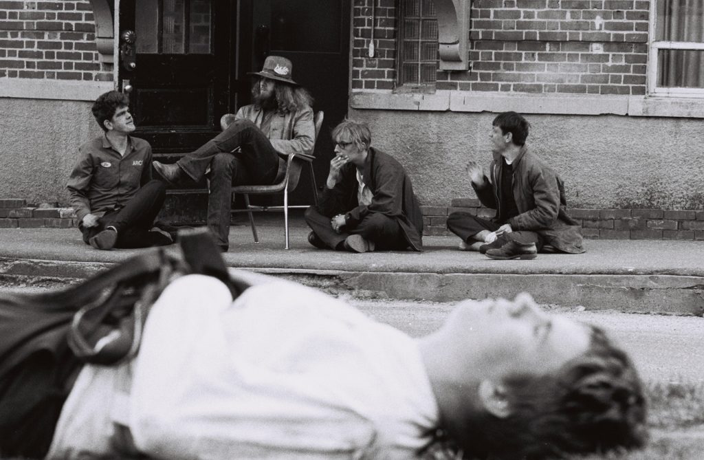 Black-and-white photograph of four people gathered on the front steps of a brick building. Three sit cross-legged on the sidewalk while another sits in a chair in the doorway, and they appear to be talking casually. In the foreground, out of focus, a fifth person lies on the ground with their face turned upward, creating a layered composition that draws attention to both the relaxed group and the blurred figure closest to the camera.
