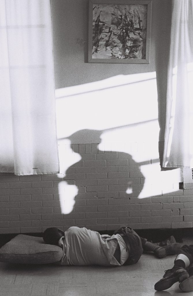 Black-and-white photograph of a person lying on their side on a floor cushion beneath a window with curtains. Sunlight casts the shadow of another person across the wall above them, and part of another person's legs are visible at the edge of the frame.