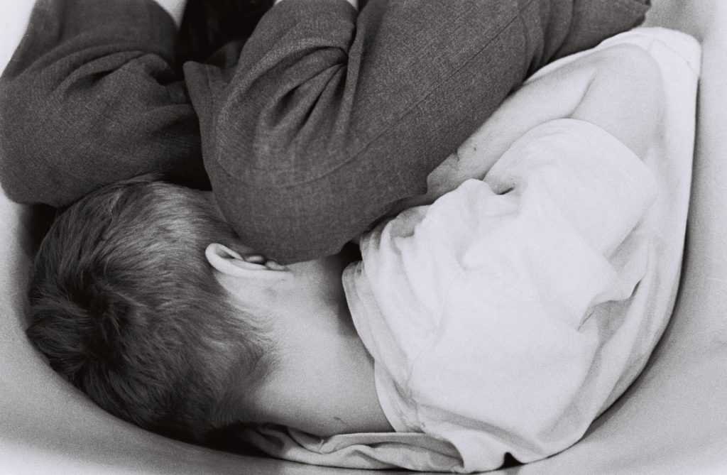 Black-and-white close-up photograph of a person lying curled on their side with their knees drawn to their chest. Their face is turned away from the camera as they rest on a pillow.