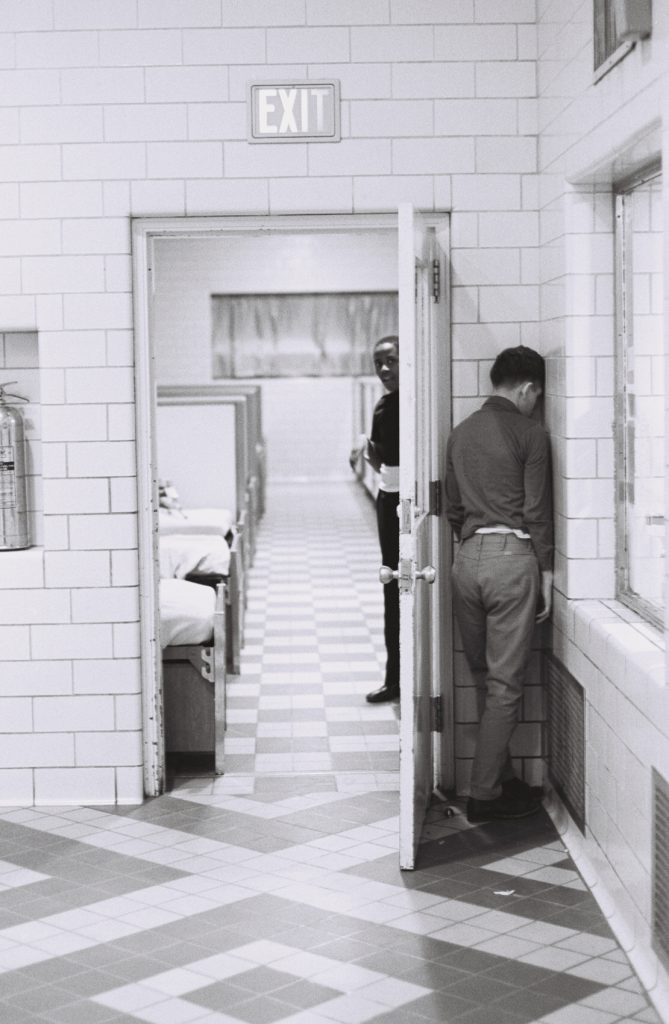 Black-and-white photograph of a tiled hallway outside a dormitory room. One person stands facing the corner with their forehead against the wall, while another looks through a partially open doorway into a room lined with beds. An EXIT sign hangs above the doorway.