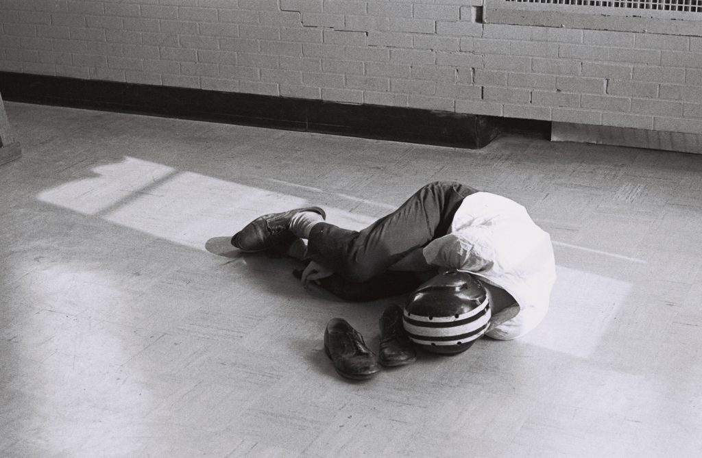 Black-and-white photograph of a person lying curled on the floor in a patch of sunlight. A striped helmet and a pair of shoes rest beside them, with a brick wall in the background.