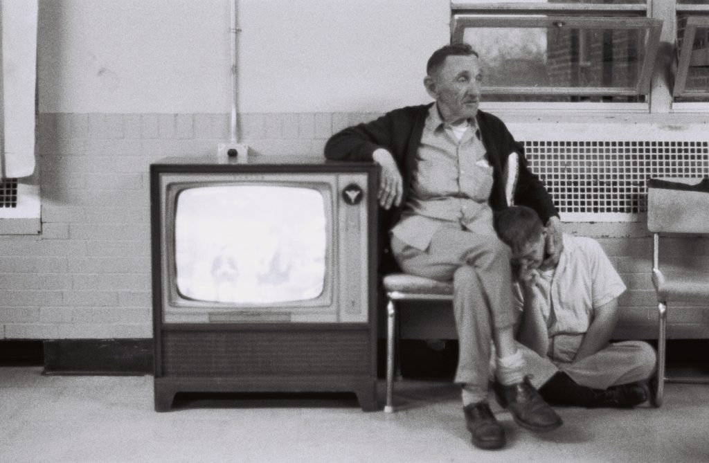 A resident sits on the floor leaning over and resting his head on another seated elderly resident who smiles.