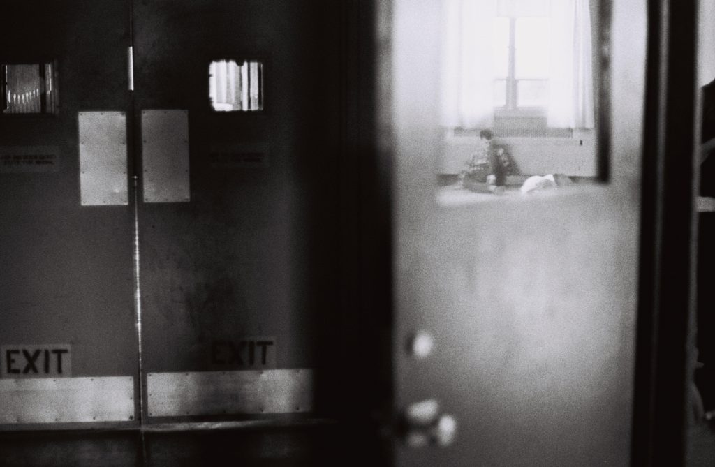 Black-and-white photograph looking through institutional doors toward a resident sitting alone on the floor in a room beyond.