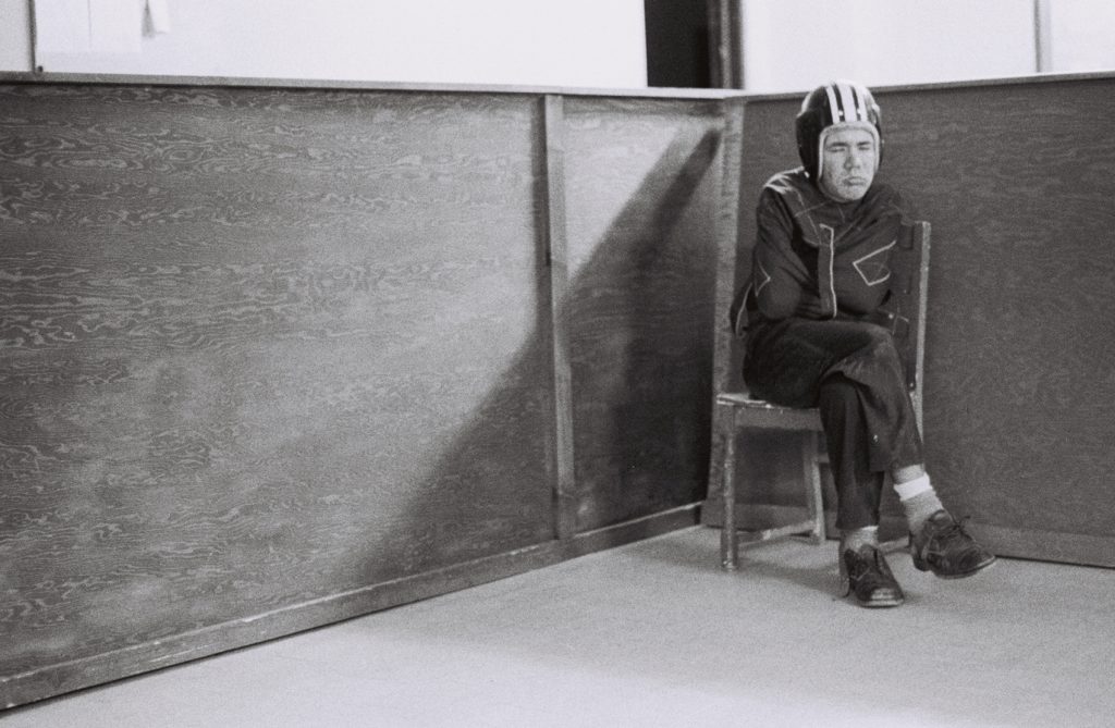 Resident wearing a protective helmet sits on a wooden chair in the corner of a room enclosed by wooden partitions.