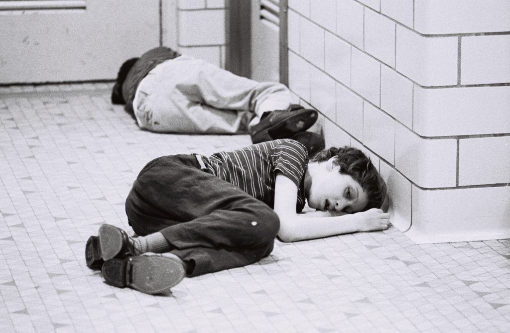 Two residents lie on a tiled hallway floor near a wall, with one in the foreground and another farther down the corridor.