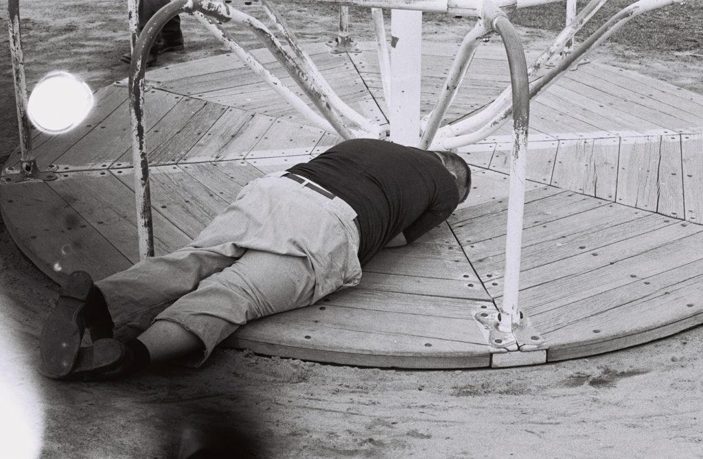 Resident lies beneath a stationary playground merry-go-round outdoors with head and arms tucked underneath himself, faced down.