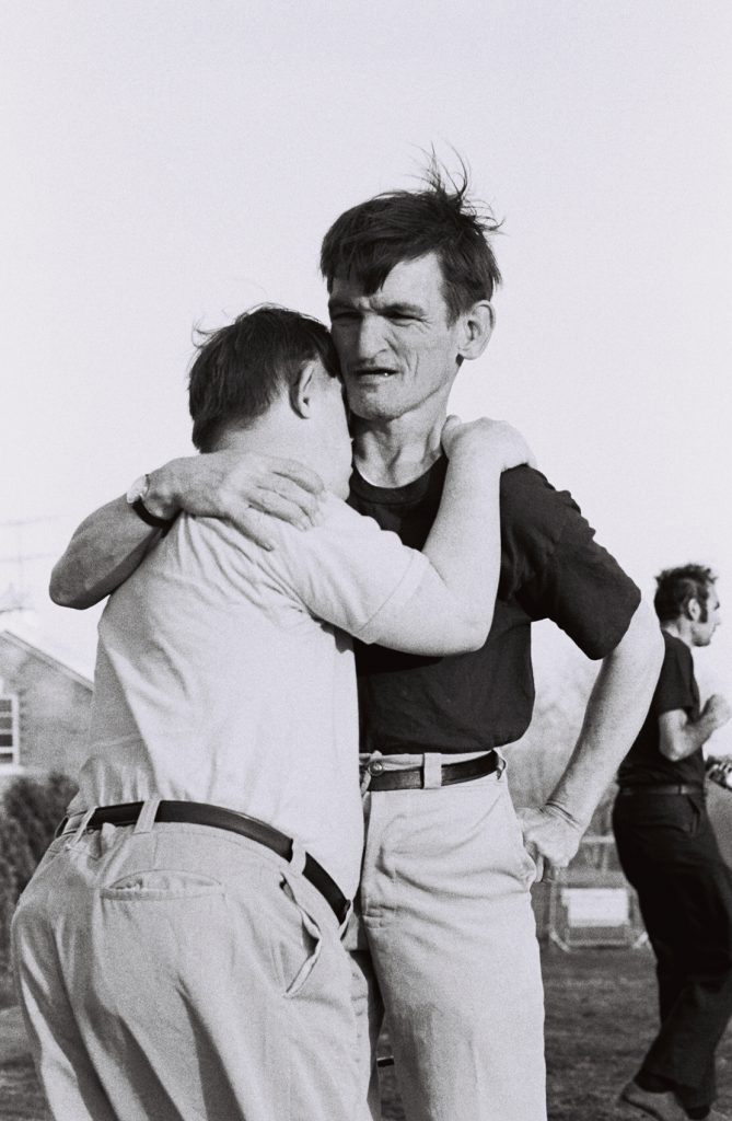 Two residents embrace outdoors while standing together; another person walks in the background.