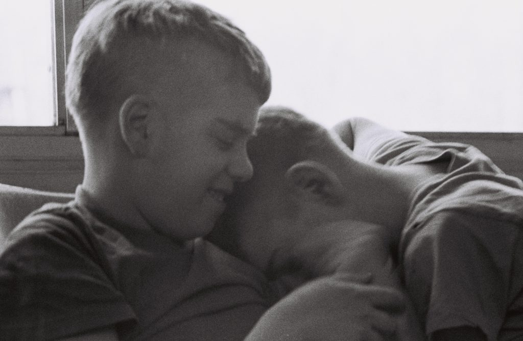 Two residents sit closely together by a window, with one resting their head against the other's shoulder and chest.