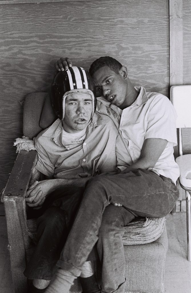 Two residents sit together in a worn upholstered chair, with one wearing a protective helmet and the other sitting on his lap leaning against him.