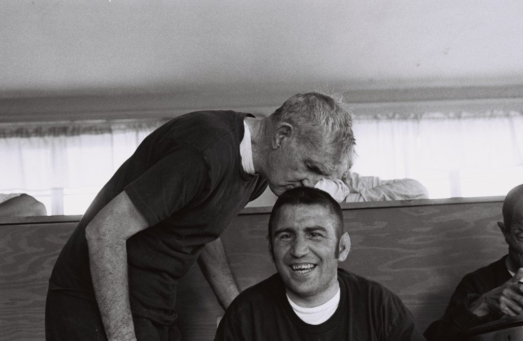 A resident sits smiling at the camera while an older resident leans down to plant a kiss atop his head.