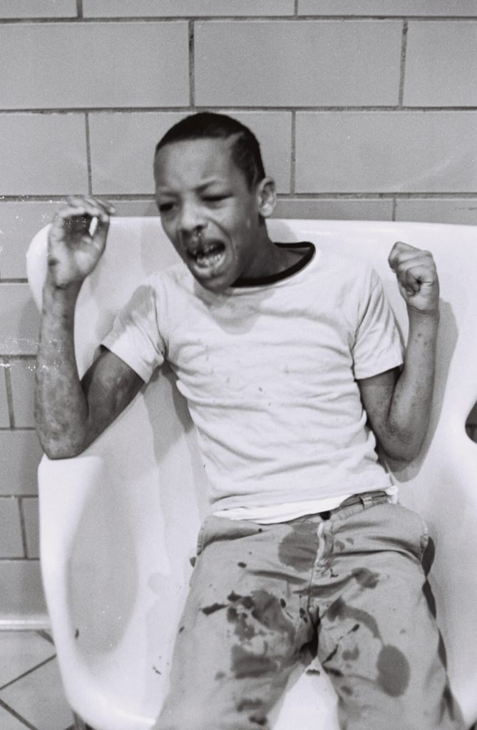 Black-and-white photograph of a resident seated in a bathtub against a tiled wall, with arms raised and mouth open. Dark stains are visible on the resident's shirt and pants.