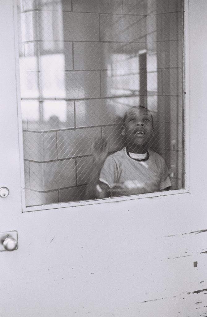 Black-and-white photograph of a child looking upward through the wired-glass window of a closed door, with one hand raised against the glass.