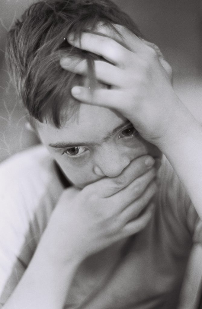 Black-and-white close-up portrait of a person resting one hand on their head and the other over their mouth while looking directly toward the camera.