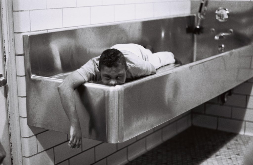 Black-and-white photograph of a person lying face down across the edge of a large institutional bathtub, with one arm hanging over the side in a tiled bathing room.