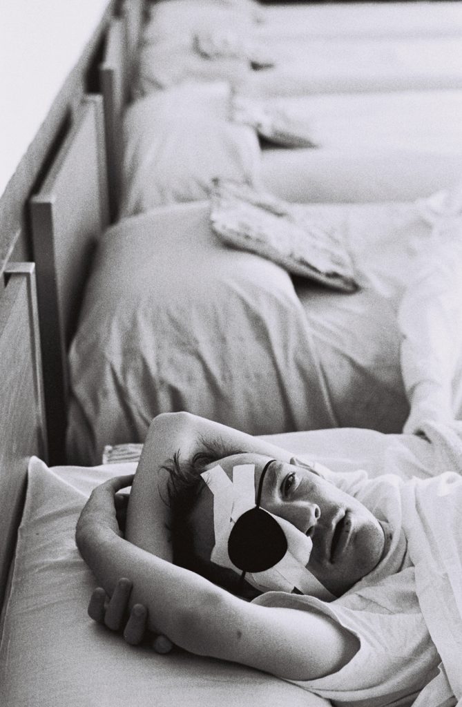 Black-and-white photograph of a resident lying in bed beneath a blanket, facing the camera with an eye covered by a bandage and protective eye patch. Additional beds extend into the background.