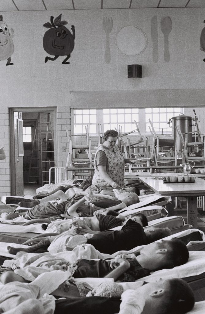 Black-and-white photograph of multiple residents lying on cots arranged in rows inside a large room while a staff member stands among them. Tables, stacked chairs, and colorful themed wall decorations are visible in the background.