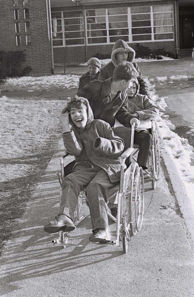 Black-and-white photograph of several residents outdoors on a winter day. Two wheelchairs are aligned on a sidewalk while other residents stand or walk nearby in front of an institutional building.
