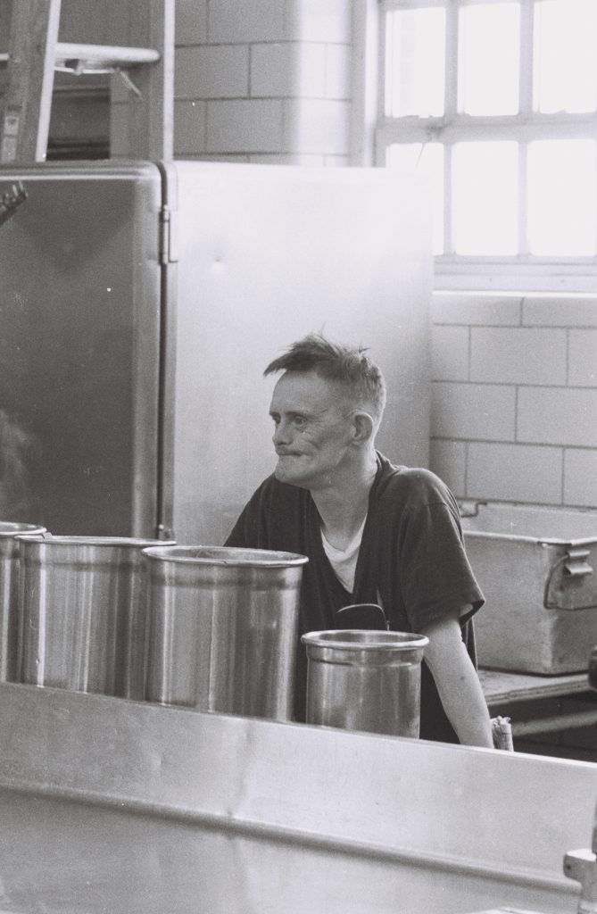 Black-and-white photograph of a resident standing behind large metal cooking pots in an institutional kitchen, looking to the side. Large windows and industrial kitchen equipment are visible in the background.