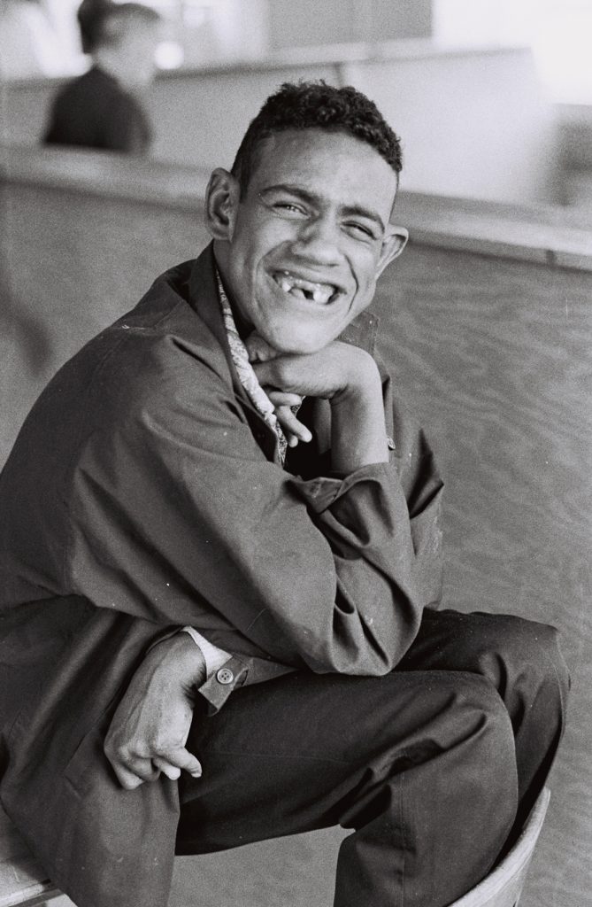 Black-and-white photograph of a smiling resident seated indoors with chin resting on one hand, looking toward the camera. A blurred figure appears in the background.