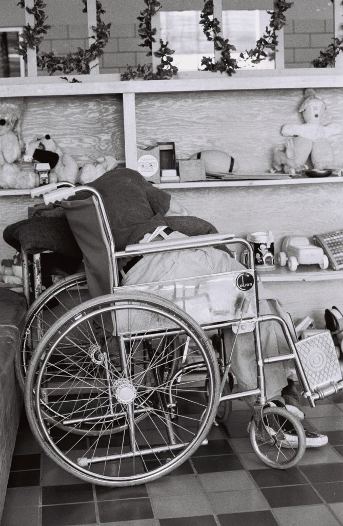 Black-and-white photograph of a person seated in a wheelchair with their upper body bent to the side, facing a shelf filled with stuffed animals, toys, and other objects in a recreation area.