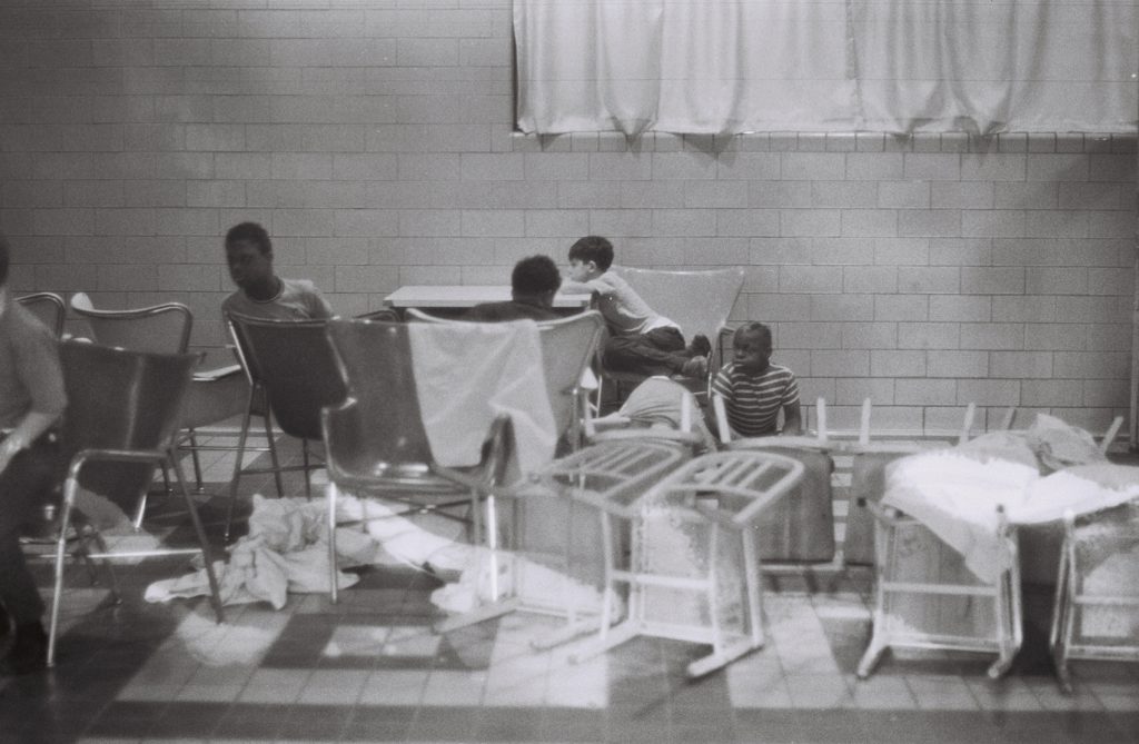 Black-and-white photo of a group of boys in a tiled institutional room, seated in metal-frame chairs arranged haphazardly. Several chairs with white linens sit in the foreground; a boy climbs over the back of a chair while others sit nearby.