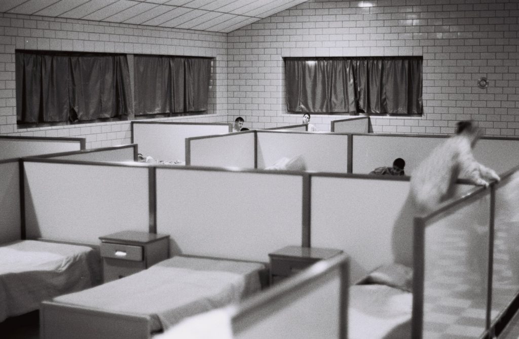 Black-and-white photo of a dormitory-style room with rows of low partition dividers separating beds, each with a nightstand. Several boys peek over the partitions from adjoining bed areas; curtained windows line the tiled wall in the background.