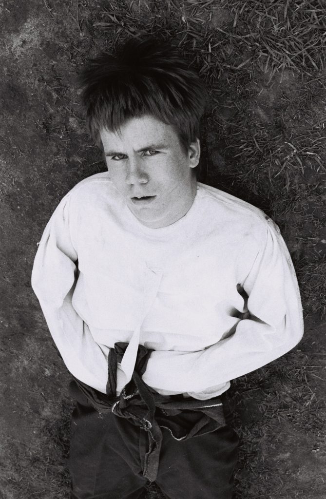 Black-and-white photo of a boy lying on the ground outdoors, wearing a white shirt tied at the waist with a dark sash, looking up toward the camera.