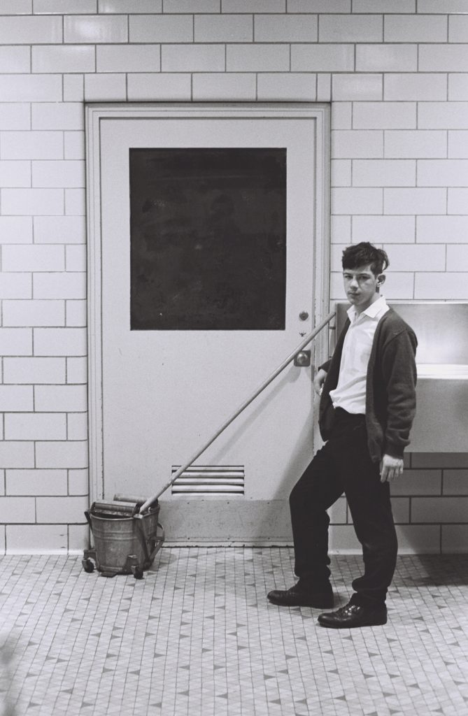 A person stands beside a mop bucket and mop in a tiled hallway, leaning against the mop handle in front of a closed door. A drinking fountain is mounted on the wall to the right.