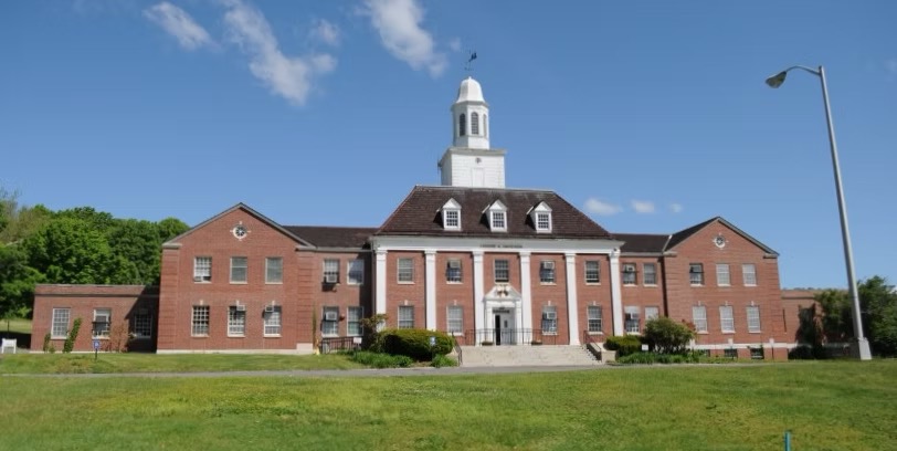 Front view of the Southbury Training School's main administration building, a large red-brick institutional building with a white-columned entrance and central cupola, set behind a grassy lawn.